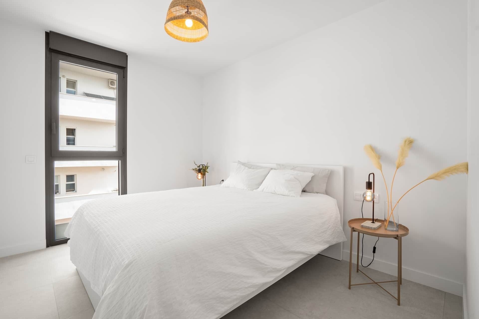 A minimalist bedroom with a large bed, a small side table, and a window with a view of buildings.