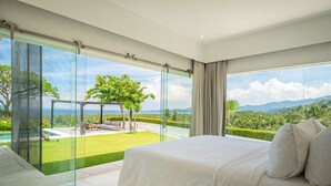 Ein modernes Schlafzimmer mit einem großen Bett, einer Glastür, die auf einen Balkon führt, und Blick auf einen üppigen Garten sowie Berge.