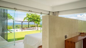 Ein modernes Schlafzimmer mit einem großen Fenster, einem Bett, einem Holztisch und Blick auf einen üppigen Garten und den Ozean.