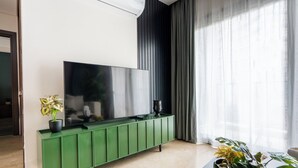 Ein modernes Wohnzimmer mit einem großen Flachbildfernseher, der an einer grünen Kommode befestigt ist, einer Topfpflanze links und einer dekorativen Schale rechts.