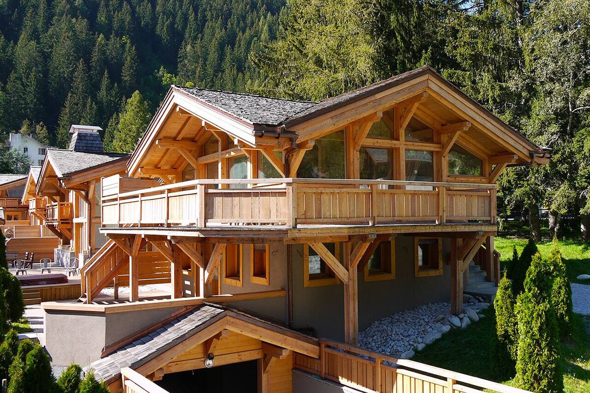 A wooden chalet with a balcony, surrounded by a forested mountain backdrop.
