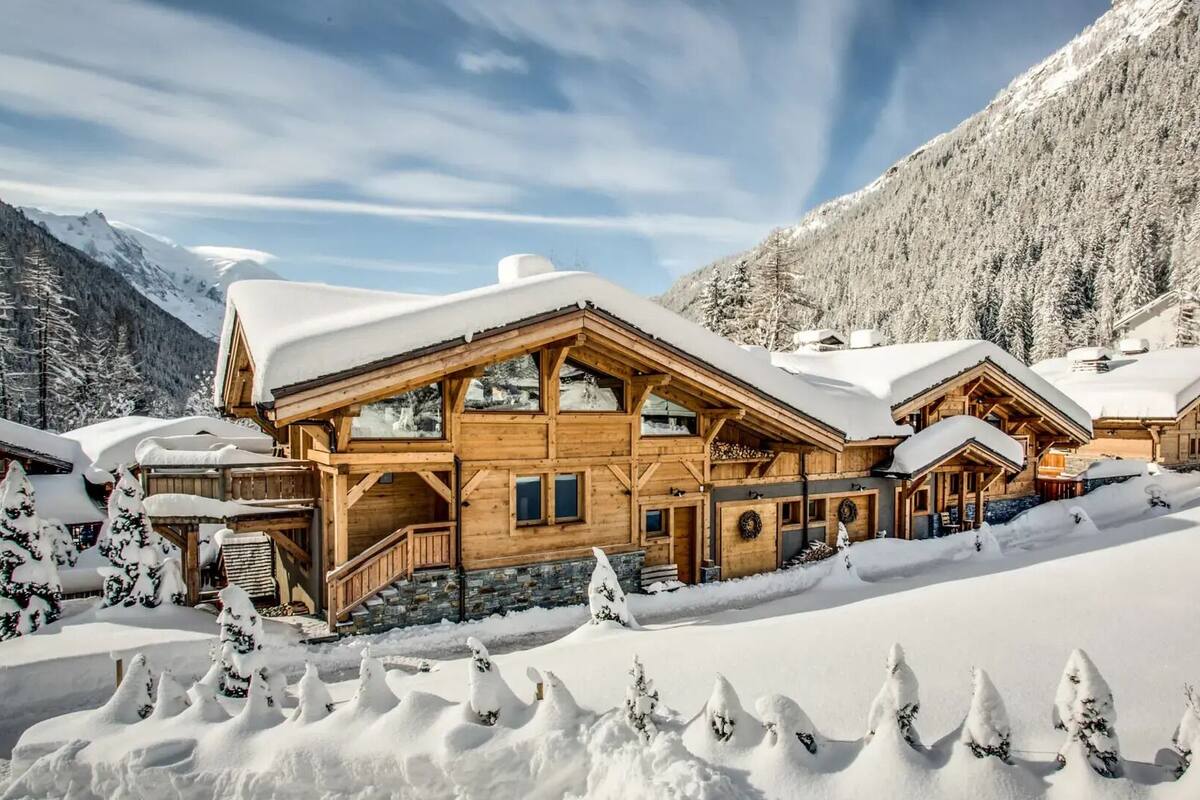 A wooden chalet with a large balcony, surrounded by snow-covered trees and mountains.