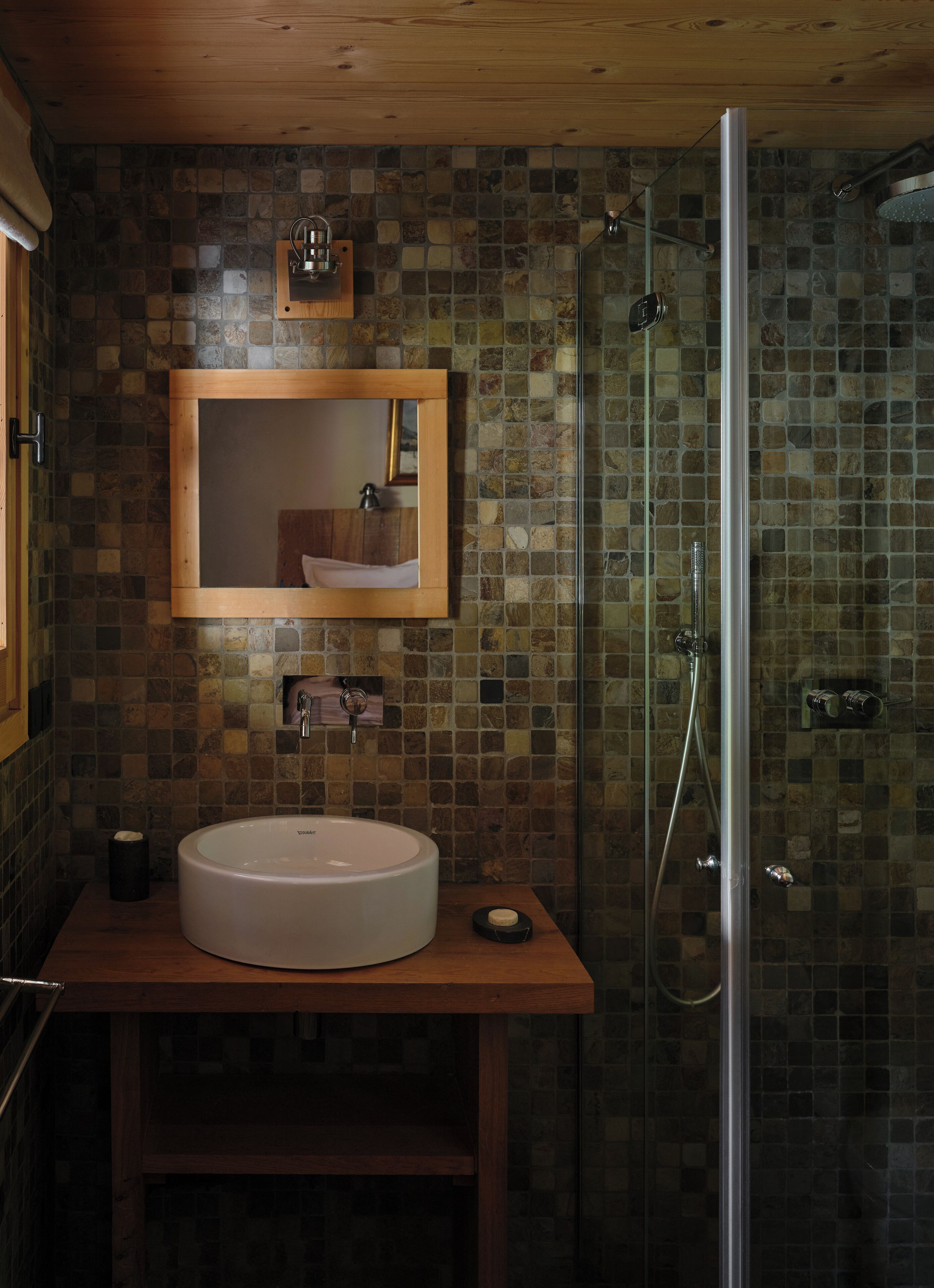 A bathroom with a stone mosaic wall, a wooden vanity, a large sink, a mirror, and a glass-enclosed shower.