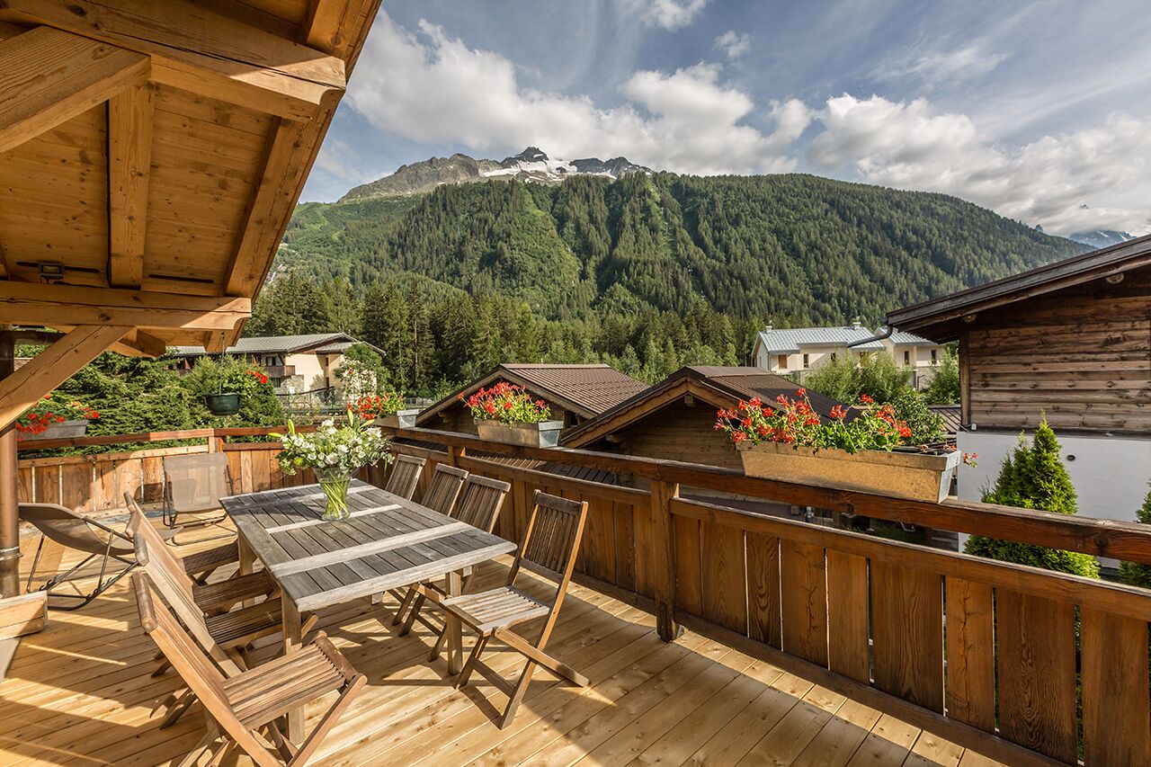 A wooden deck with a table and chairs, overlooking a mountain and a lake.