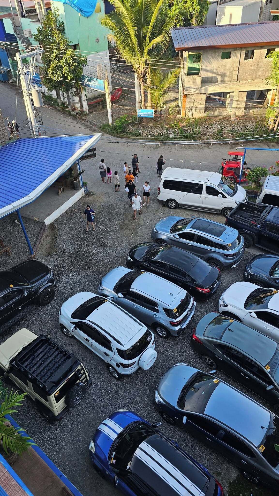 Un estacionamiento con varios autos y un autobús, rodeado de edificios y una palmera.