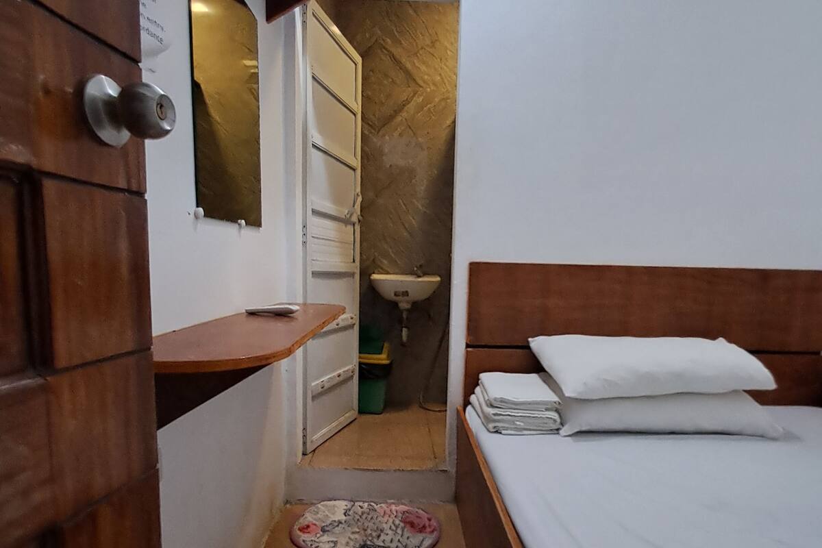 A narrow room with a wooden wardrobe, a small bedside table, a bed with white bedding, and a bathroom with a sink and mirror.