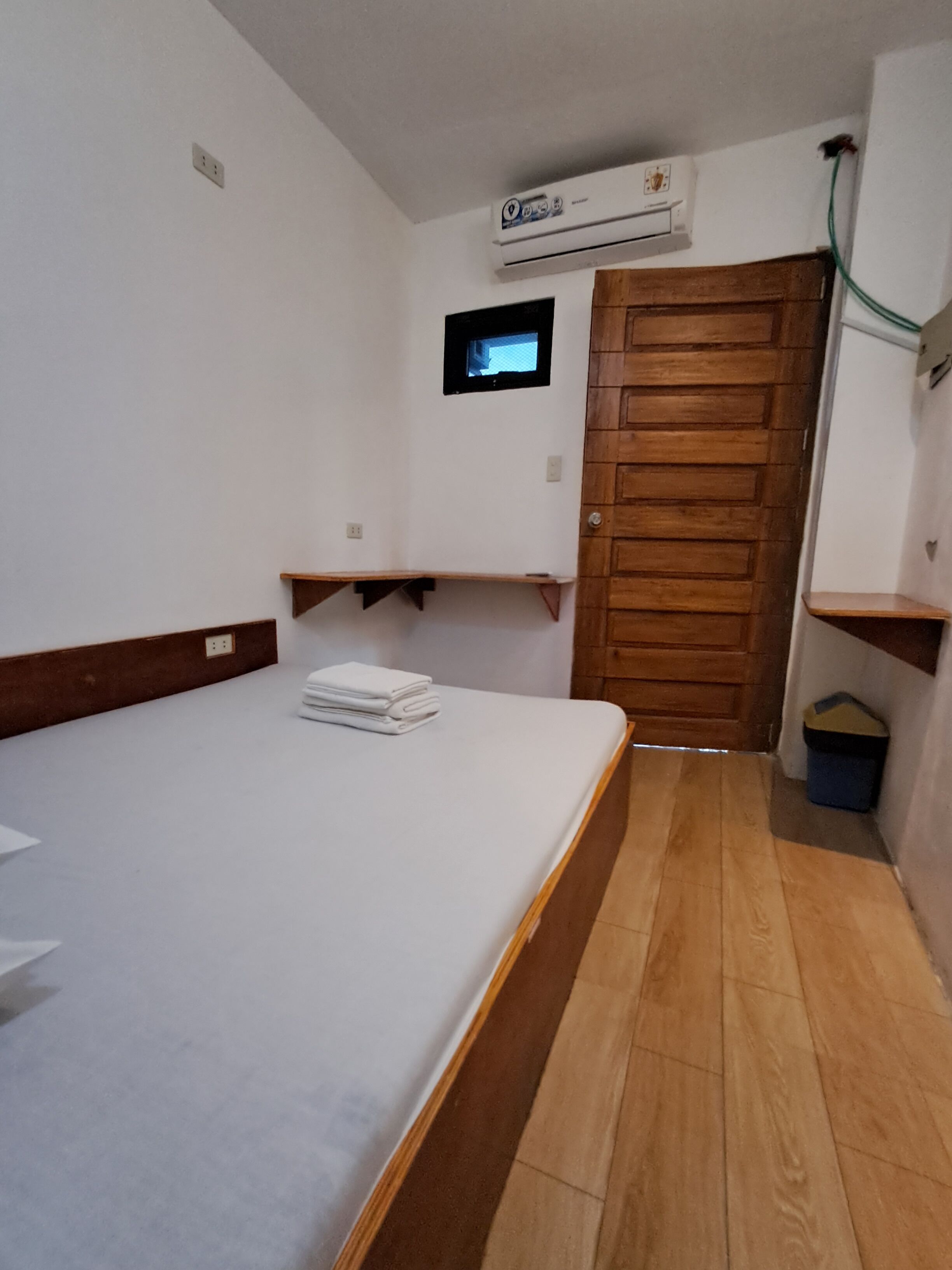 A small, clean room with a bed, a wooden door, a small window, and a wall-mounted air conditioner.