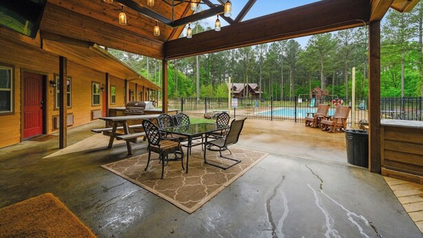 A covered patio with a dining table and chairs, a pool, and a view of the surrounding forest. - Big Timber Lodge (Broken Bow)