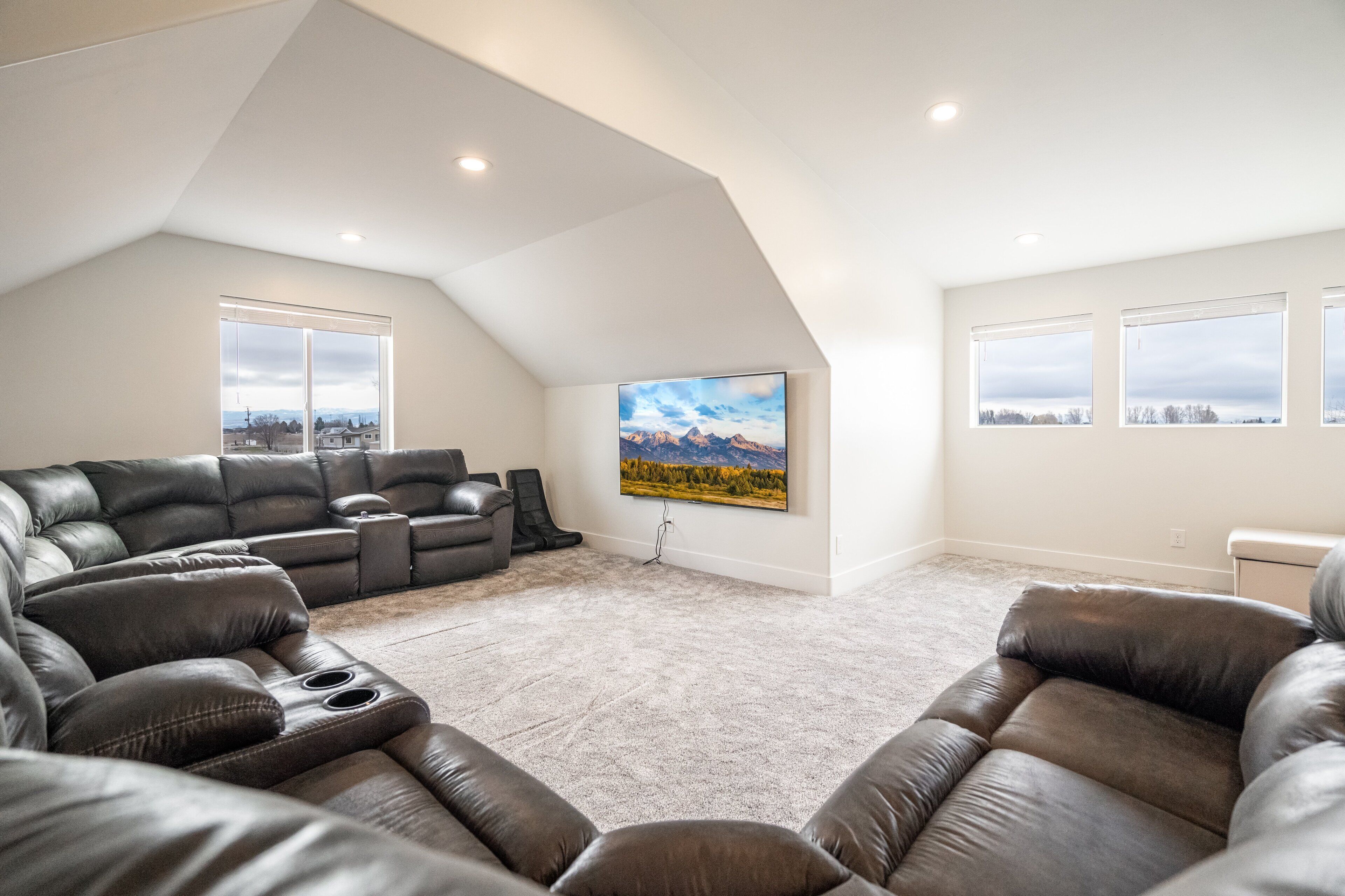 A spacious living room with a large sectional sofa, a flat-screen TV mounted on the wall, and a view of the outdoors through large windows.