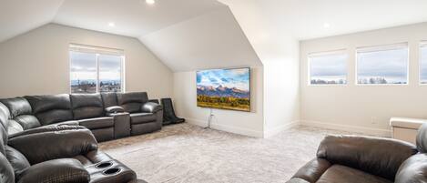 A spacious living room with a large sectional sofa, a flat-screen TV mounted on the wall, and a view of the outdoors through large windows.