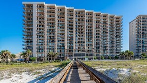 Un hotel de altura con varios balcones, un paseo marítimo de madera que conduce a la entrada y un cielo azul despejado.