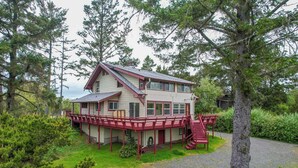 A two-story house with a red exterior and a balcony, surrounded by trees. - Multi-level home, large wrap around deck, stunning views of the Necanicum River. (Seaside)