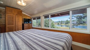 A bedroom with a large bed, wooden cabinets, and a window with a view of a lake and buildings. - Multi-level home, large wrap around deck, stunning views of the Necanicum River. (Seaside)