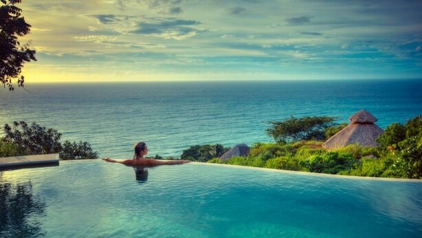 En person som kopplar av i en vacker, turkosblå infinitypool med utsikt över havet och byggnader med halmtak.