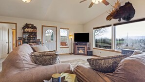 A cozy living room with a fireplace, comfortable seating, a television, and a large window offering a view. - Pet Friendly Home with Fishing Access and Fire Pit (Kalispell)