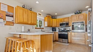 A kitchen with wooden cabinets, a central island with stools, stainless steel appliances, and a corkboard with photos. - Pet Friendly Home with Fishing Access and Fire Pit (Kalispell)
