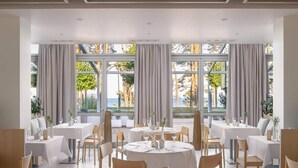 Un restaurant avec des tables et des chaises rondes, de grandes fenêtres avec des rideaux et une vue sur l’extérieur.
