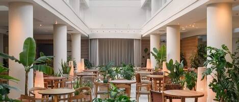 A spacious indoor area with multiple round tables and chairs, surrounded by large potted plants, and a high ceiling with skylights.