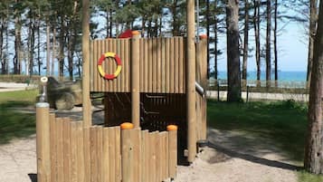 Puinen leikkialue, jossa on liukumäki ja kiipeilyelementtejä, ja jonka ympärillä on puita ja hiekkaista maaperää.