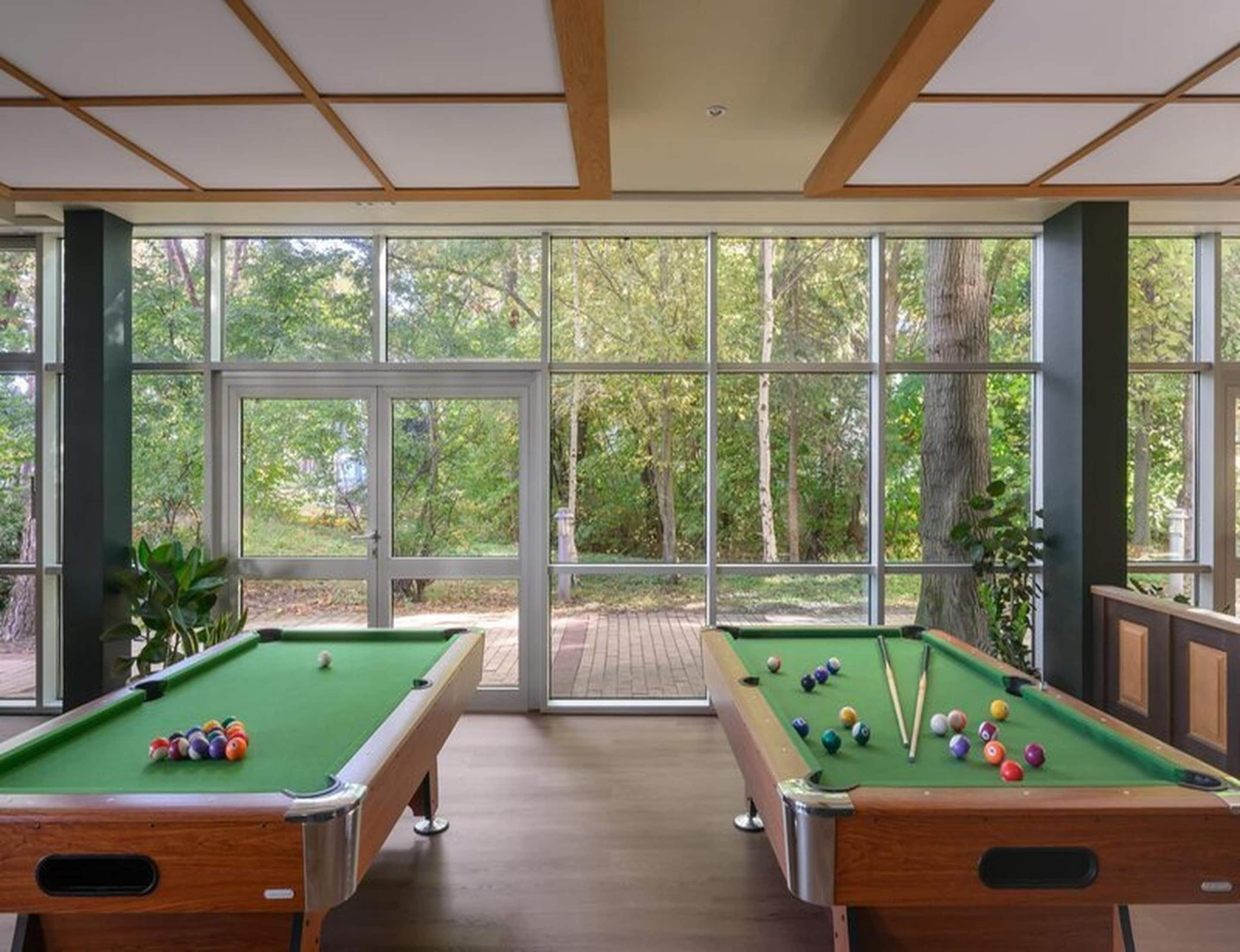 A pool table with balls, a second table, and a large window with a view of trees.
