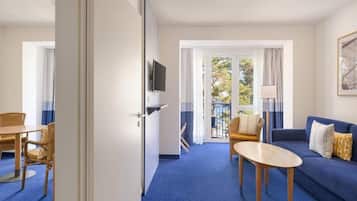 Ein Hotelzimmer mit Essbereich, einer Couch, einem an der Wand montierten Fernseher und einem Balkon mit Vorhängen.