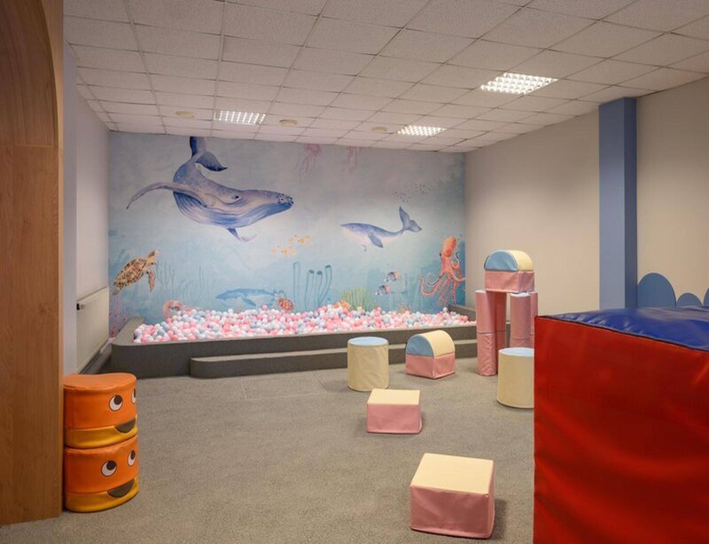 Zona de juegos infantiles con un mural de criaturas marinas, piscina de pelotas y mobiliario acolchado.