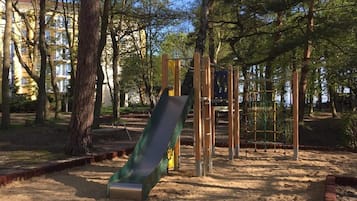 Ein Spielplatz mit Rutsche, Klettergerüsten und Sandkasten, umgeben von Bäumen und Gebäuden.