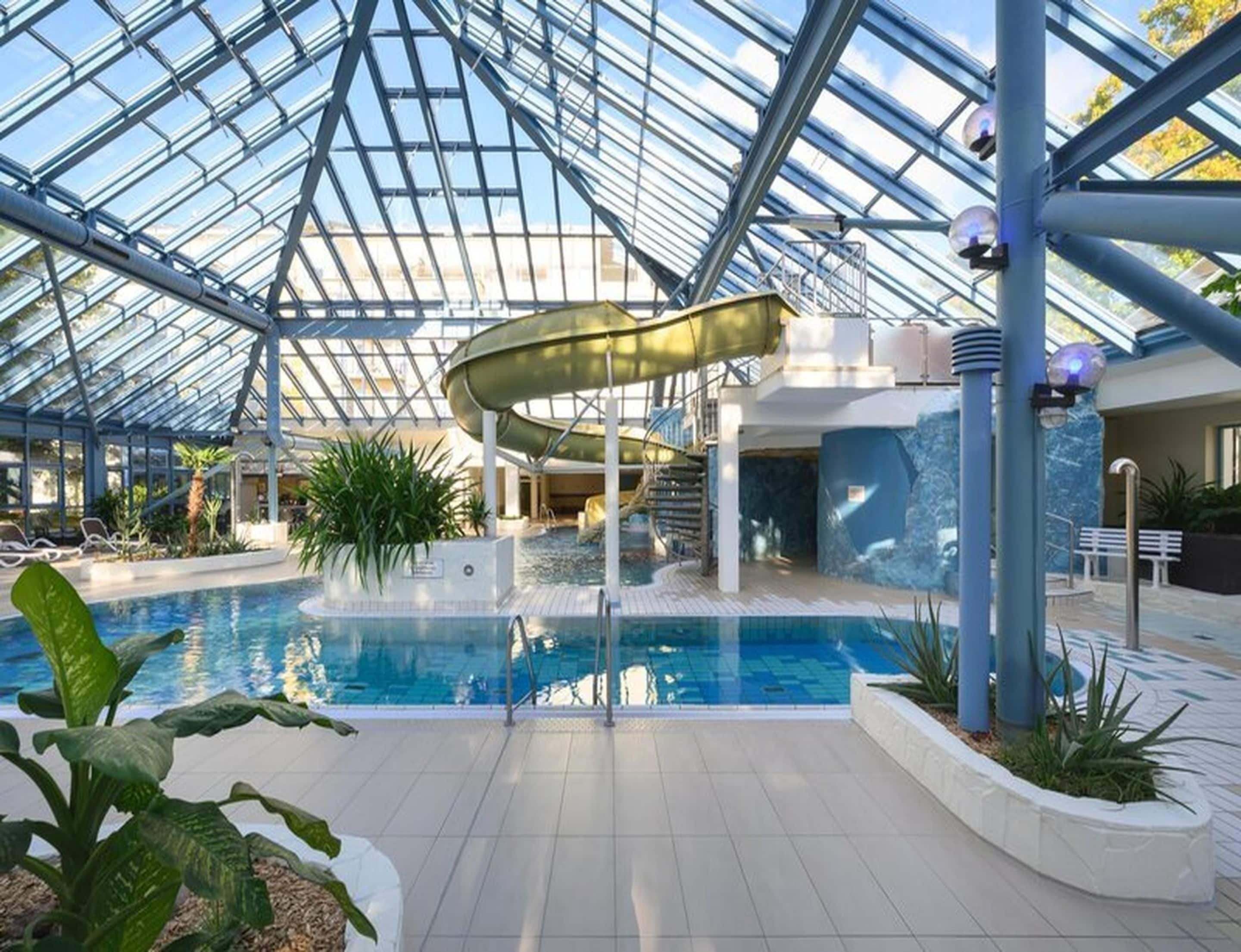 Ein Hallenbad mit Glasdach, einer Rutsche und einer Sitzgelegenheit mit Pflanzen.