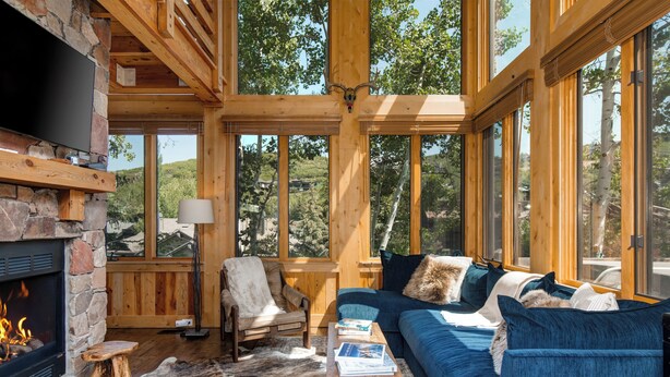 Interior acogedor de una cabaña con chimenea de piedra, muebles de madera y grandes ventanas que ofrecen vistas al bosque circundante.