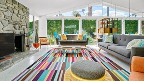 A modern living room with a stone fireplace, large windows, and a colorful striped rug. - Twin Palms: Mid-Century Modern with Heated Pool (Palm Springs)