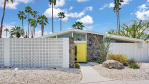 A modern house with a yellow door, surrounded by a white fence and palm trees. - Twin Palms: Mid-Century Modern with Heated Pool (Palm Springs)