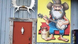 Un bâtiment rustique avec une porte rouge, une fresque représentant un homme barbu jouant du banjo, et une figurine de cow-boy sur le toit.