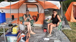 Two people seated at a table under a tent, with camping gear and supplies visible. - Walk to Rock The South Tent/Car Camping (Cullman)