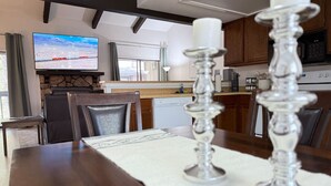 Un coin repas avec une table en bois, deux chandeliers, un téléviseur et une cuisine en arrière-plan.