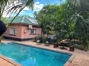 Piscina com espreguiçadeiras e uma mesa, rodeada por vegetação exuberante e um edifício com telhado verde.
