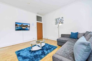 A modern living room with a grey sofa, a glass coffee table, a blue rug, a painting, and a floor lamp.