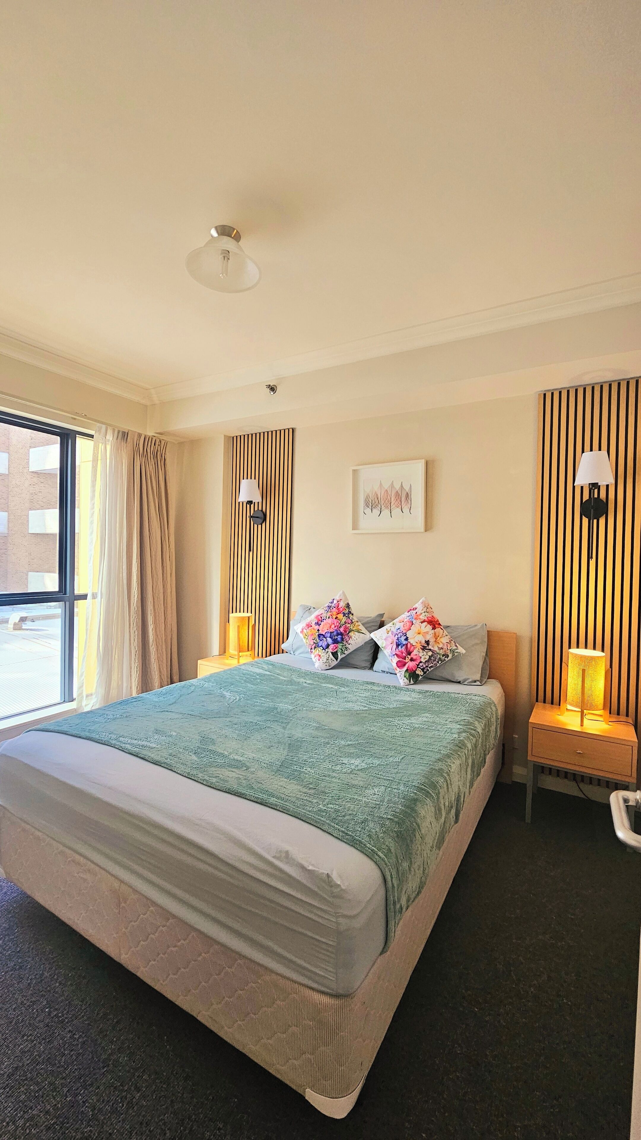 A hotel room with a large bed, two bedside lamps, a window with curtains, a wall-mounted artwork, and a ceiling light.