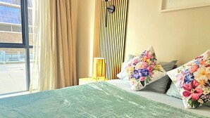 Ein Hotelzimmer mit einem großen Bett, floralen Kissen, einer Stehlampe und einem Fenster mit Vorhängen.