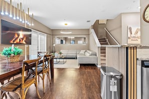 A modern living room with a large flat-screen TV, wooden dining table, and comfortable seating. A staircase leads to an upper level, and a trash can is visible. - NEW 4Bedrooms At Lucaya/Close to Disney (Kissimmee)