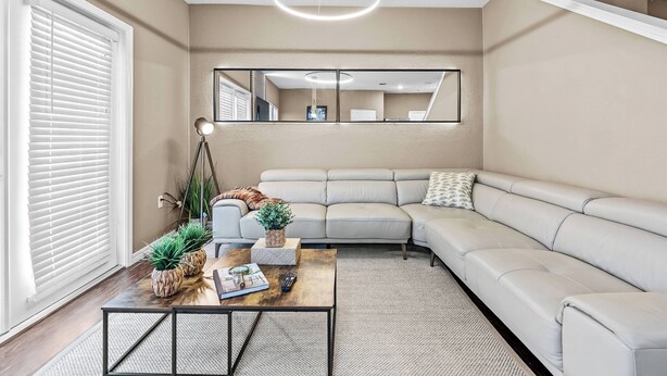 A modern living room with a beige sofa, a coffee table with decorative items, a large mirror, and a ceiling light fixture. - 4BR house with pool near Disney area (Kissimmee)