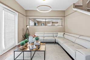 Un salon moderne avec un canapé beige, une table basse agrémentée d’objets décoratifs, un grand miroir et un luminaire suspendu au plafond.