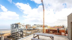 A rooftop terrace with a view of the city and beach, featuring a crane, a picnic table, and a parking area.