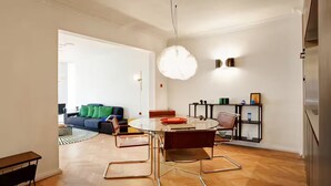 A modern dining area with a glass table and metal chairs, a sofa with green cushions, and a bookshelf with decorative items.