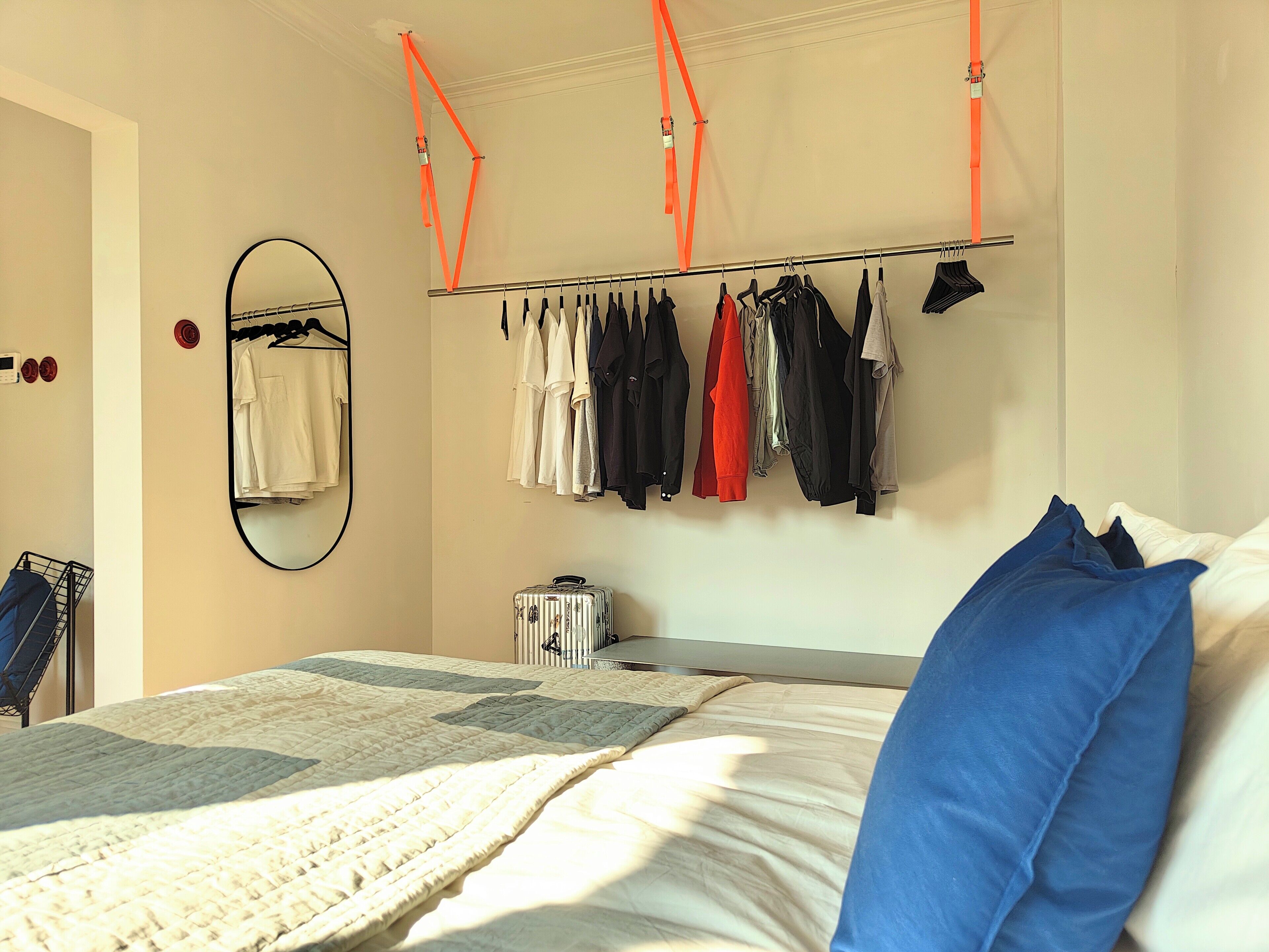 A bedroom with a bed, a hanging clothes rack, a suitcase, and a mirror on the wall.
