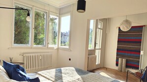 A bedroom with a large bed, a radiator, a window with a view of greenery, and a hanging light fixture.