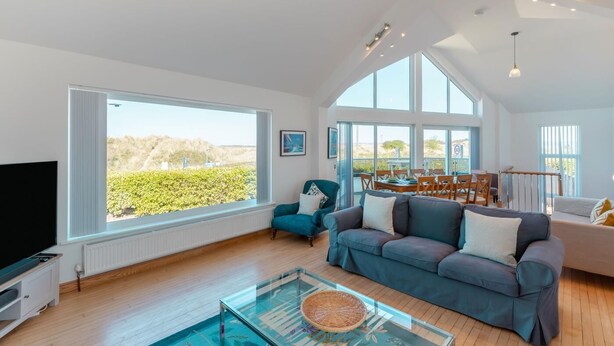 A spacious living room with a large window, a glass coffee table, a sofa, and a dining area with chairs. - 4 Bed in Instow (oc-i31748) (Instow)
