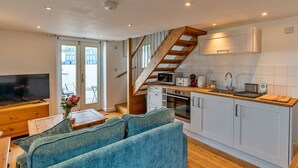 A modern kitchen with white cabinets, a wooden staircase, and a dining area with a blue sofa. - Cottage in Porthleven Near Beach & Harbour (Helston)