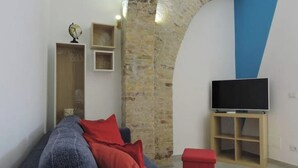Un salon avec un canapé bleu, des coussins rouges, un téléviseur posé sur un meuble en bois et un mur en briques.