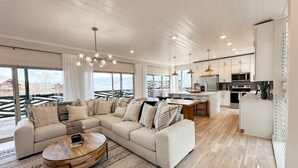 A spacious living area with a large L-shaped sectional sofa, a wooden coffee table, and a dining table with chairs. The room features a high ceiling with recessed lighting, large windows offering a view of the outdoors, and a modern kitchen with white cabinetry and stainless steel appliances. - Smoky Mountain Views / Luxury Escape (Gatlinburg)