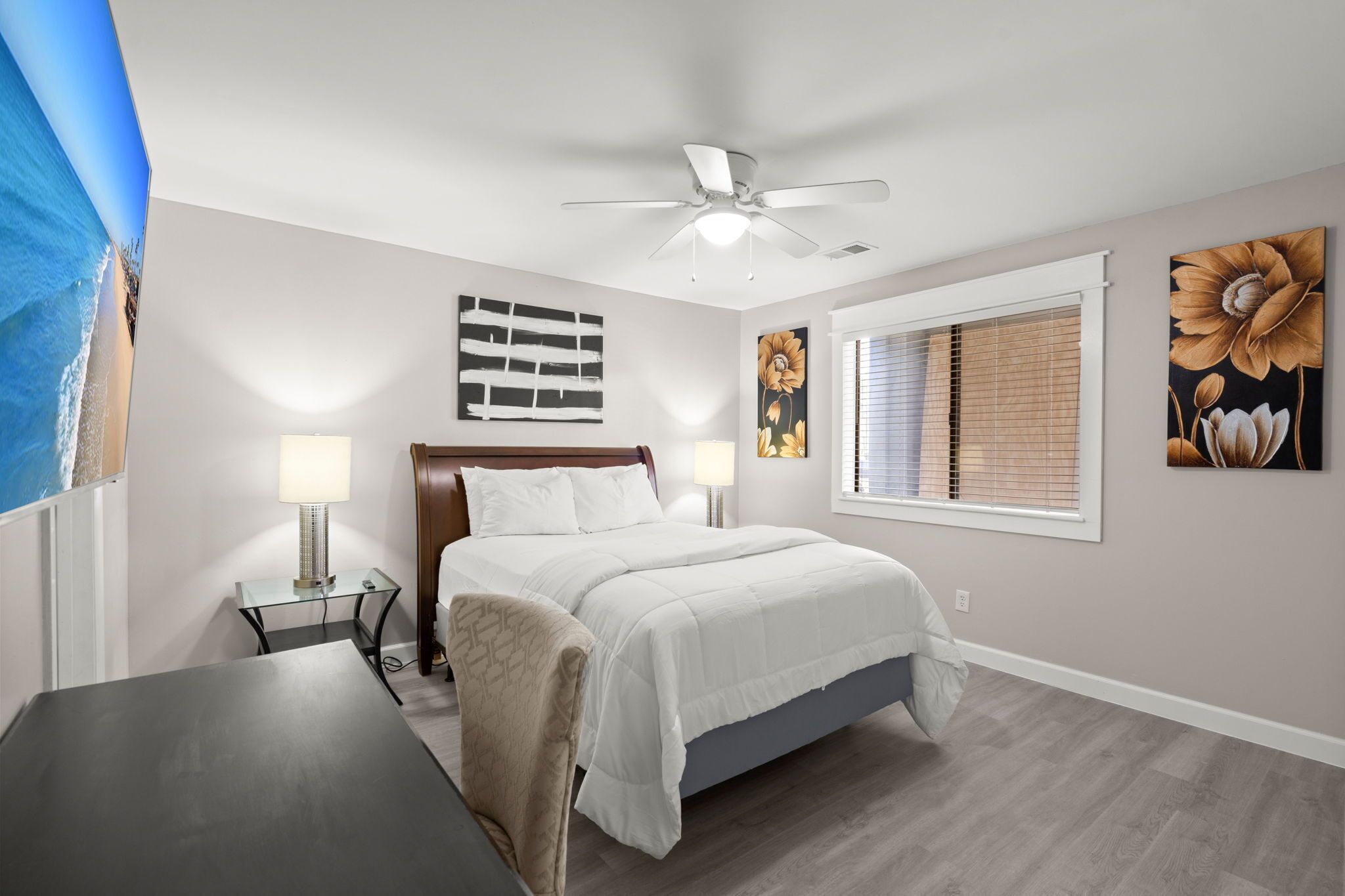 A bedroom with a bed, bedside lamps, a ceiling fan, a window with blinds, and artwork on the walls.